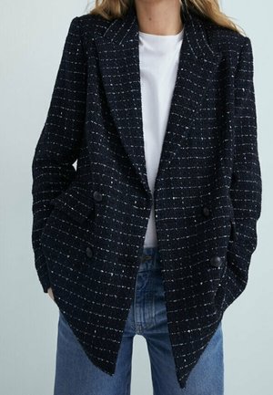 Femme portant un blazer croisé à carreaux bleu marine sur un t-shirt blanc et un jean bleu, les mains dans les poches du blazer.