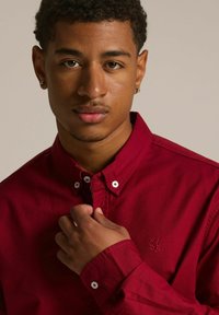 Czerwona koszula z kołnierzykiem wykonana z bawełny, z zapięciem na guziki oraz haftowanym logo. Posiada białe guziki oraz detale mankietów. Gładka tekstura.