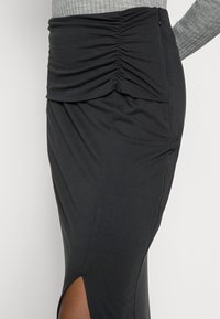 Jupe noire ajustée avec une taille froncée, détail de fente sur le côté et tissu lisse et extensible. Fermeture éclair à l'arrière.