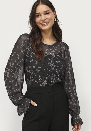 Zwarte doorschijnende blouse met bloemenpatroon, lange mouwen en ruches aan de mouwen. Gecombineerd met hoge zwarte broeken. Lachende model.