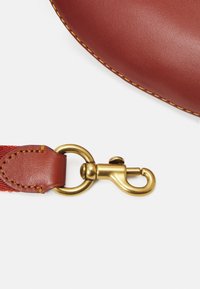 Dettaglio di una borsa in pelle con corpo arrotondato di colore rosso intenso, dotata di un moschettone girevole in ottone e di una tracolla intrecciata per una connessione sicura.