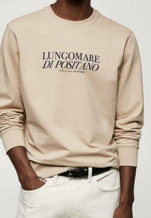 Beige langærmet skjorte med teksten "LUNGOMARE DI POSITANO Italia, Costa Amalfitana", båret med hvide bukser og sort bælte.