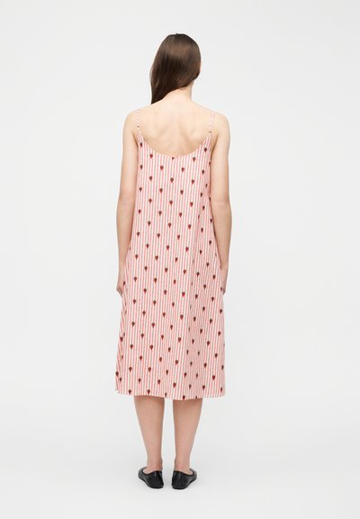 Γυναίκα που στέκεται με την πλάτη στραμμένη προς την κάμερα, φοράει φόρεμα με ροζ και λευκές ρίγες και μικρά σχέδια φράουλας, καθώς και μαύρα παπούτσια χωρίς κορδόνια.