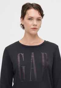 Sudadera gris oscura con cuello redondo, que presenta un gran logotipo brillante de "GAP" en tonos oscuros en la parte frontal. Textura de algodón suave.