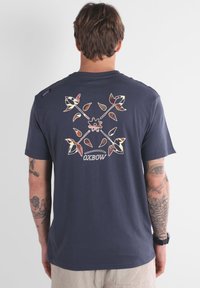 T-shirt bleu marine foncé avec un design graphique floral au dos, mettant en avant des accents crème, rose et vert. Tissu doux, manches courtes, coupe décontractée.
