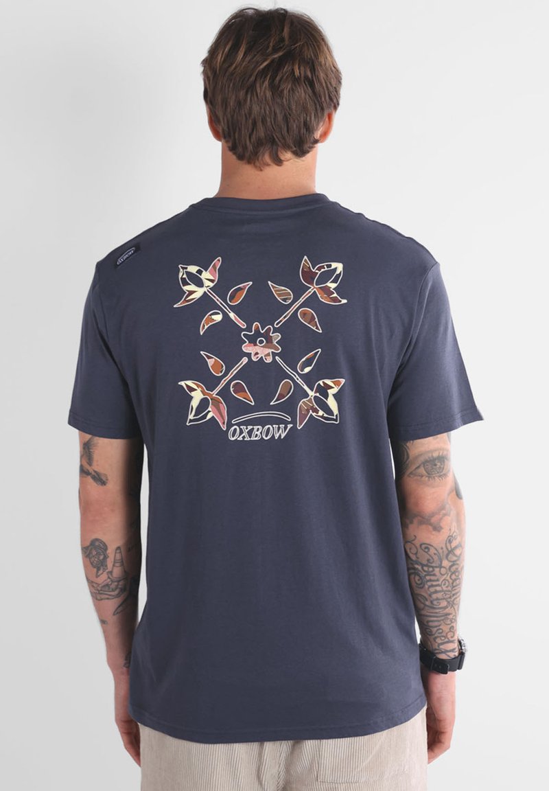 T-shirt bleu marine foncé avec un design graphique floral au dos, mettant en avant des accents crème, rose et vert. Tissu doux, manches courtes, coupe décontractée.