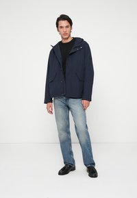 Tengerészkék vízálló dzseki kapucnival, két elülső zsebbel és patentos zárással, világoskék farmernadrággal és fekete cipővel párosítva.