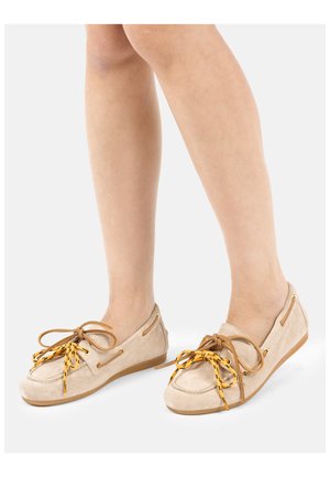 Nogi w beżowych zamszowych mokasynach z żółtymi i brązowymi kokardkami, stojące na białej powierzchni.