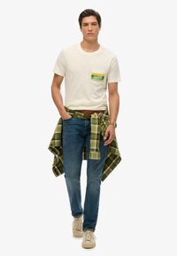 Camiseta blanca de manga corta con un bolsillo verde y amarillo, jeans azules y una camisa a cuadros verde atada a la cintura. Zapatillas beige y cinturón marrón.