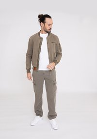 Chaqueta bomber ligera de color verde oliva con cierre de cremallera, puños de canalé y detalles en los bolsillos, combinada con pantalones cargo y zapatillas blancas.