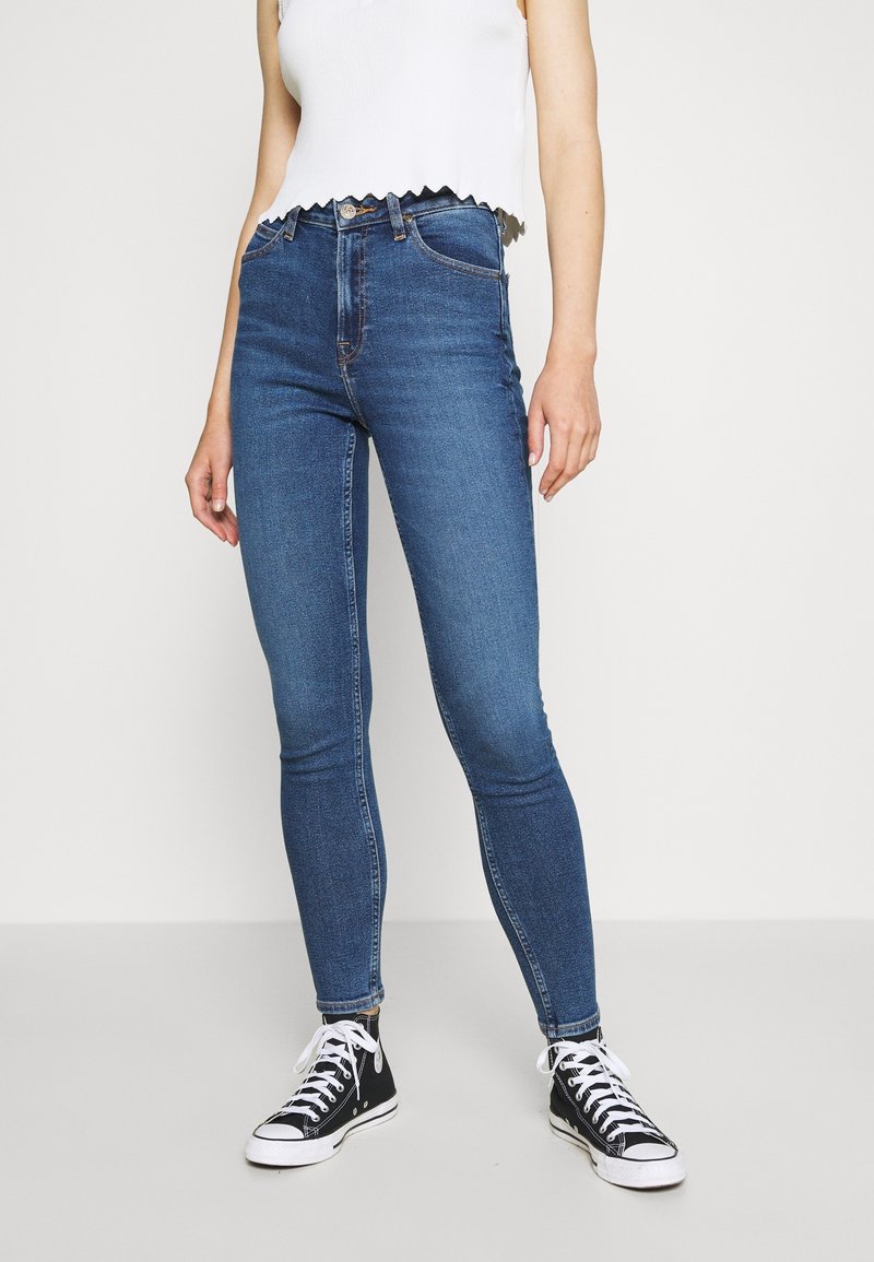 Jeans skinny azules fabricados en denim, con una cintura alta, diseño de cinco bolsillos y detalles desgastados, combinados con zapatillas negras.