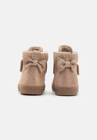 Tan suede ankle boots with two decorative bows on the back, soft interior lining, and textured rubber sole. Lightweight and short-cut design.