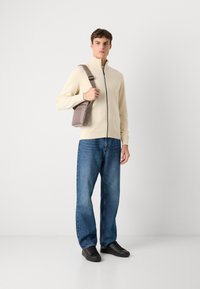 Un pull zippé beige clair, un jean bleu et des baskets noires. Un petit sac marron est porté en bandoulière. Design simple, tenue décontractée.
