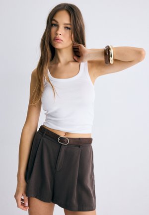 Nuori nainen pukeutuneena valkoiseen toppiin ja korkeavyötäröisiin ruskeisiin shortseihin vyöllä, poseeraa yhdellä kädellä taivutettuna ja rannekoruilla ranteessa.