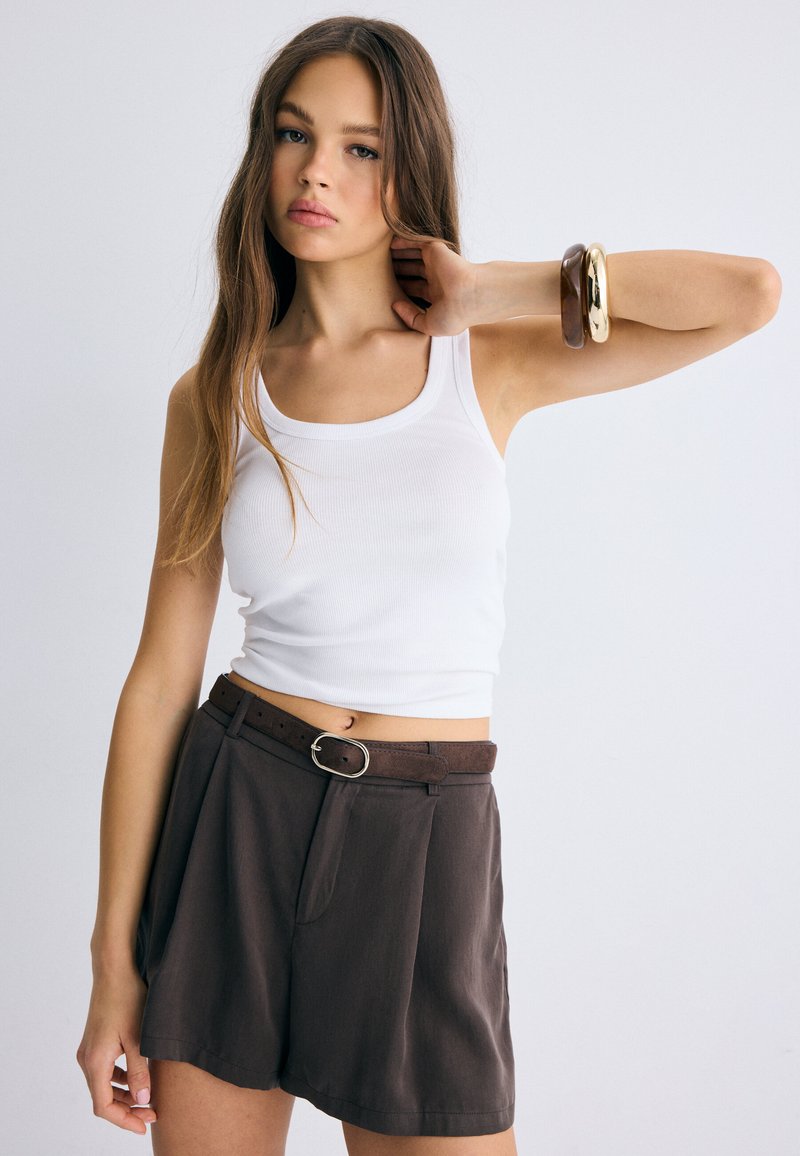 Young woman wearing white tank top and high-waisted brown shorts with belt, posing with one arm bent and bracelets on wrist.
