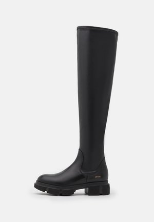 Botte noire montante jusqu’au genou avec semelle épaisse, dessus lisse et petit logo doré près du talon, vue de côté sur fond blanc.