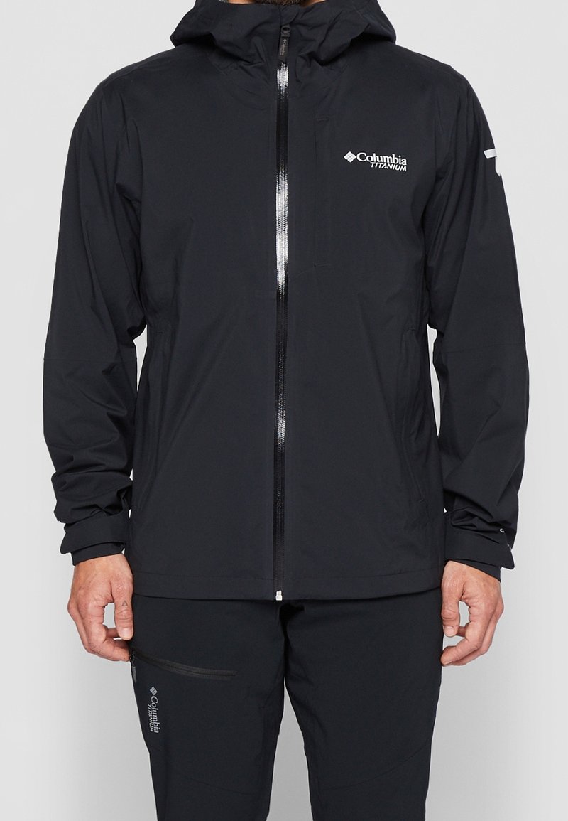 Homme portant une veste à capuche à zip intégral noire Columbia Titanium et un pantalon noir avec les logos Columbia visibles sur la poitrine et la cuisse.
