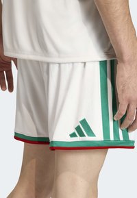 Bijeli sportski šortsevi s zelenim i crvenim detaljima. Imaju tri vertikalne zelene pruge i izvezeni zeleni logo s lijeve strane.