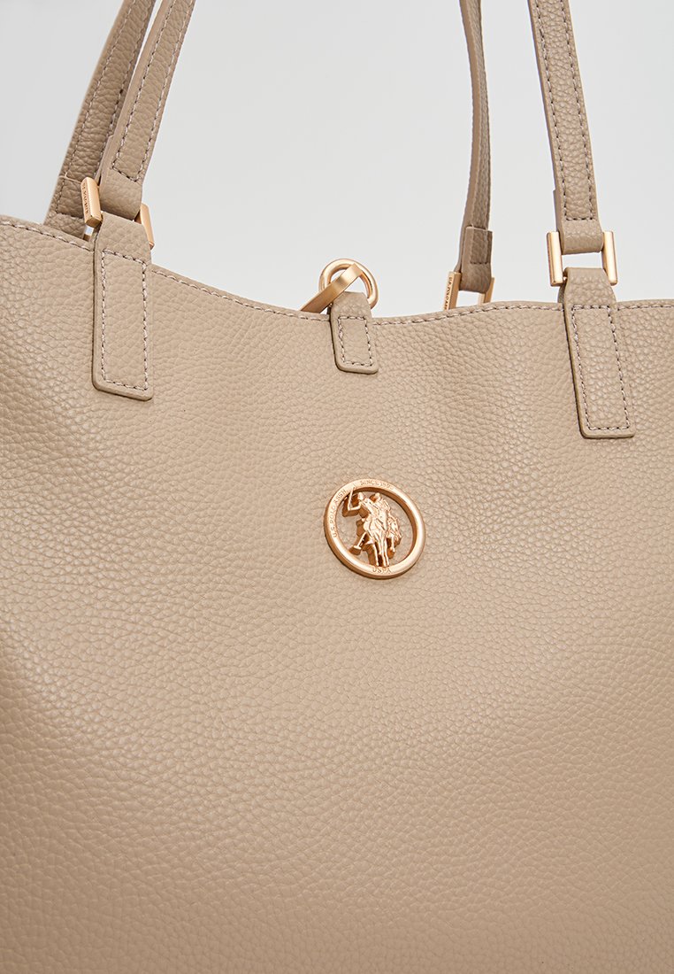 Sac fourre-tout en cuir texturé beige avec deux sangles d'épaule et un accent logo circulaire en doré. Fini lisse avec des détails de couture visibles.
