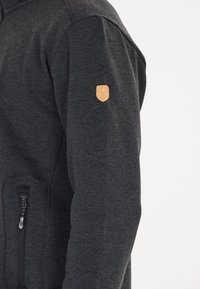 Giacca grigia scura con zip in tessuto morbido, dotata di un colletto di pelle beige sulla manica e una tasca con zip sul lato.