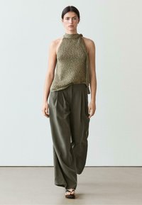 Kvinde iført ærmeløs, højhalset striktop og løse, højtaljede vidde bukser går på ensfarvet baggrund.