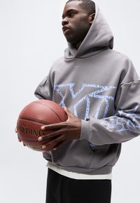 Junger Mann in grauem Hoodie, der mit beiden Händen einen Spalding-Basketball vor einem schlichten hellen Hintergrund hält.