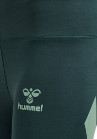 Mørkegrøn nærbillede af sportstights med hummel-logo og bier-symbol trykt nær det øvre lår.