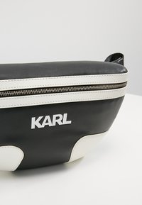 Mustavalkoinen nahkainen ristilaukku vetoketjusulkimella, jossa on rohkea logo ja tyylikäs, kaareva muotoilu. Joustava pinta.