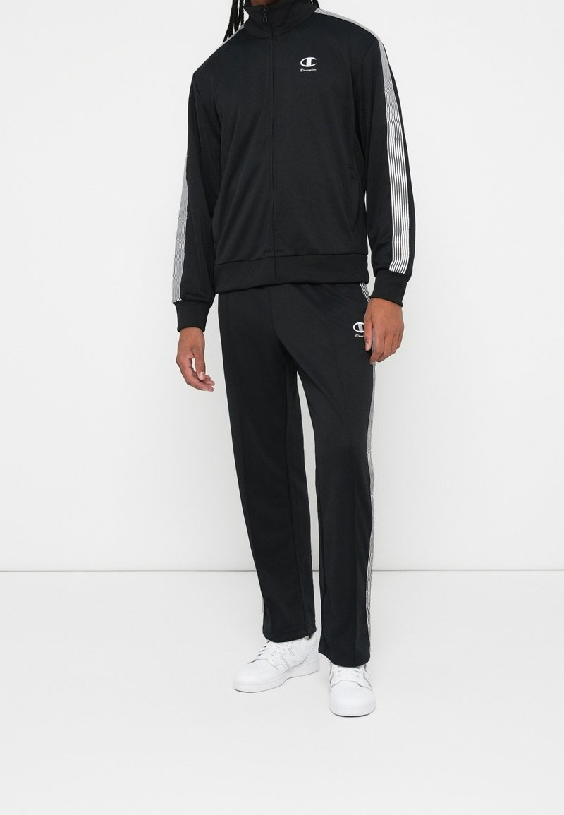 Personne portant un survêtement noir Champion avec des rayures blanches aux manches et aux jambes, assorti de baskets blanches, debout devant un fond blanc.