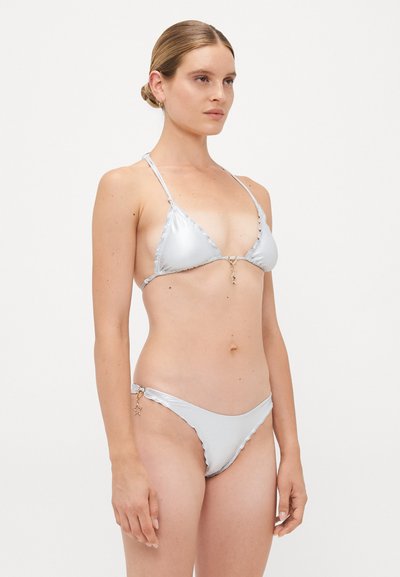 Sieviete ar bunā sakārtotiem matiem, tērpusies spīdīgā sudraba bikini ar zvaigžņu formas piekariņiem uz balta fona, vērsta nedaudz pa labi.