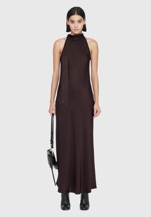 Mujer con cabello liso y oscuro que lleva un vestido largo negro sin mangas con cuello halter, sosteniendo un bolso negro y con botas negras.