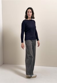 Marineblauw geribbelde longsleeve-top gecombineerd met hoge taille grijze wijde jeans en beige sneakers met blauwe accenten, tegen een neutrale achtergrond.