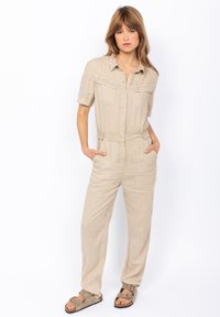 Jumpsuit in lino beige chiaro con maniche corte, chiusura con bottoni, due tasche sul petto, gambe affusolate e vita con cintura. Indossato con sandali marroni.