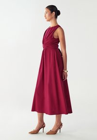 Crvena midi haljina s dizajnom preklopa sprijeda, strukirana i s lepršavom suknjom. Glatka tkanina, u kombinaciji s bež sandalama s potpeticom i šiljastim vrhom.