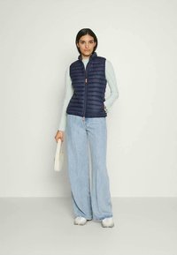 Colete acolchoado azul-marinho com costura vertical, fecho com zíper e detalhe em laranja. Usado sobre uma blusa de manga longa azul clara e combinado com jeans de perna larga.