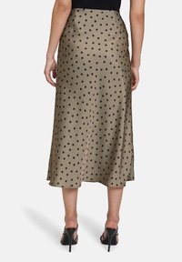 Jupe midi en satin vert olive avec de grands pois noirs. Elle présente une taille ajustée et un léger brillant. Portée avec des sandales à talons hauts noires.