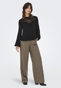 Haut noir à manches longues avec un panneau en dentelle texturé sur la poitrine, associé à un pantalon large beige et des chaussures noires.