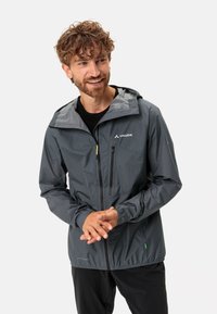 Giacca leggera grigia con cappuccio, cerniera frontale e due tasche con cerniera. Presenta dettagli a contrasto e un accento verde vicino all'orlo.
