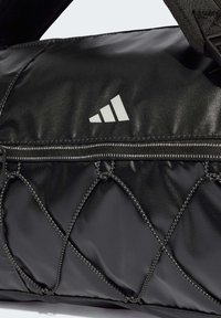 Black duffel bag with quilted design, featuring a zippered pocket, reflective logo, and sturdy nylon strap. Textured finish with contrasting stitching.
