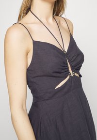Femme portant une robe sans manches sombre avec de fines bretelles, un plissé au niveau de la poitrine et une découpe sous la poitrine avec des perles décoratives sur les liens.