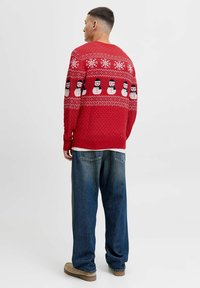 Jersey de punto rojo con un patrón de muñeco de nieve y copos de nieve blancos, con puños y dobladillo acanalados. Combinado con jeans azules de corte holgado y zapatos beige.