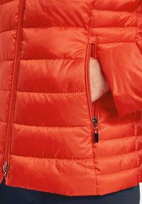 Röd puffjacka med quiltat mönster, med dragkedja framtill och sidoficka. Tyget har en slät, glänsande yta.