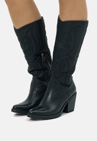 Bottes noires montantes en cuir, avec une partie inférieure en cuir et une partie supérieure en tissu ornée de motifs en spirale en relief. Talon empilé et fermeture éclair latérale.