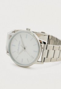 Montre-bracelet en argent Calvin Klein avec un cadran blanc texturé, des marqueurs d'heure minimalistes et un bracelet en maillons métalliques reposant sur un fond blanc.