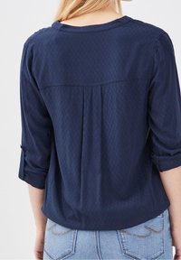 Blouse bleu marine avec un motif texturé en losange, encolure arrondie, manches trois-quarts retroussées, avec un pli au dos.