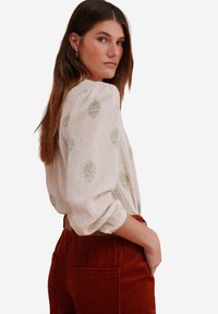 White, textured blouse with green floral patterns, paired with rust-colored corduroy pants. The blouse has billowy sleeves and gathers at the neckline.