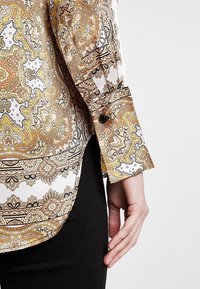 Långärmad skjorta i vitt med intrikat mönster i gult, orange och svart paisley; har en knappmanschett och rundad nederkant.