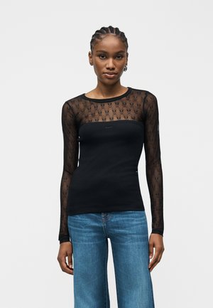 Γυναίκα που φοράει μαύρο διάφανο μπλουζάκι με μακριά μανίκια και σχεδιασμό με μοτίβο, καθώς και μπλε τζιν, στέκεται μπροστά σε απλό λευκό φόντο.