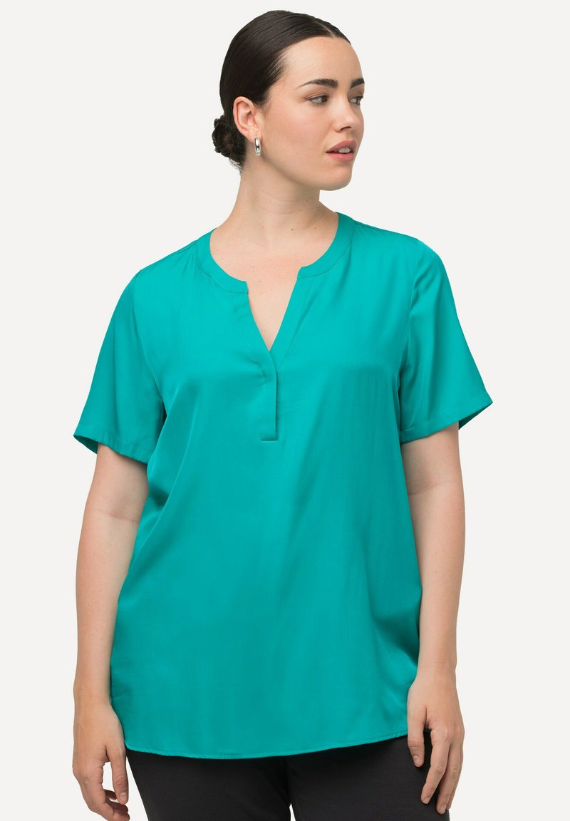 Donna con capelli scuri raccolti in uno chignon, che indossa orecchini a cerchio d'argento e una blusa azzurra a maniche corte, che guarda verso destra, su uno sfondo chiaro e semplice.