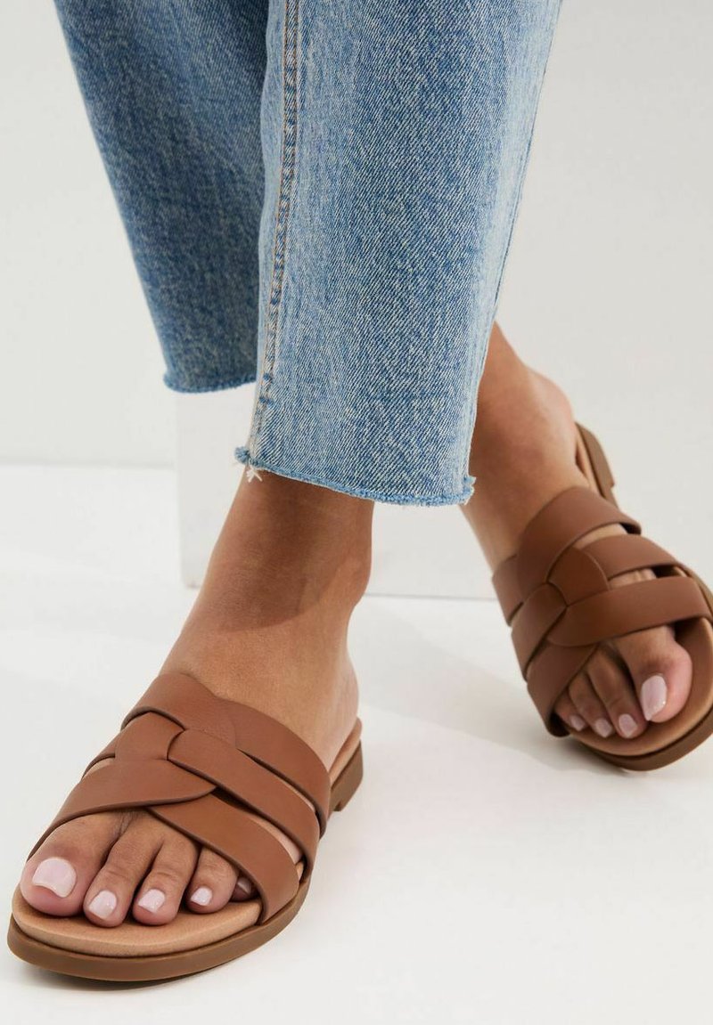 Pieds portant un jean bleu clair et des sandales plates en cuir tressé marron sur une surface blanche.